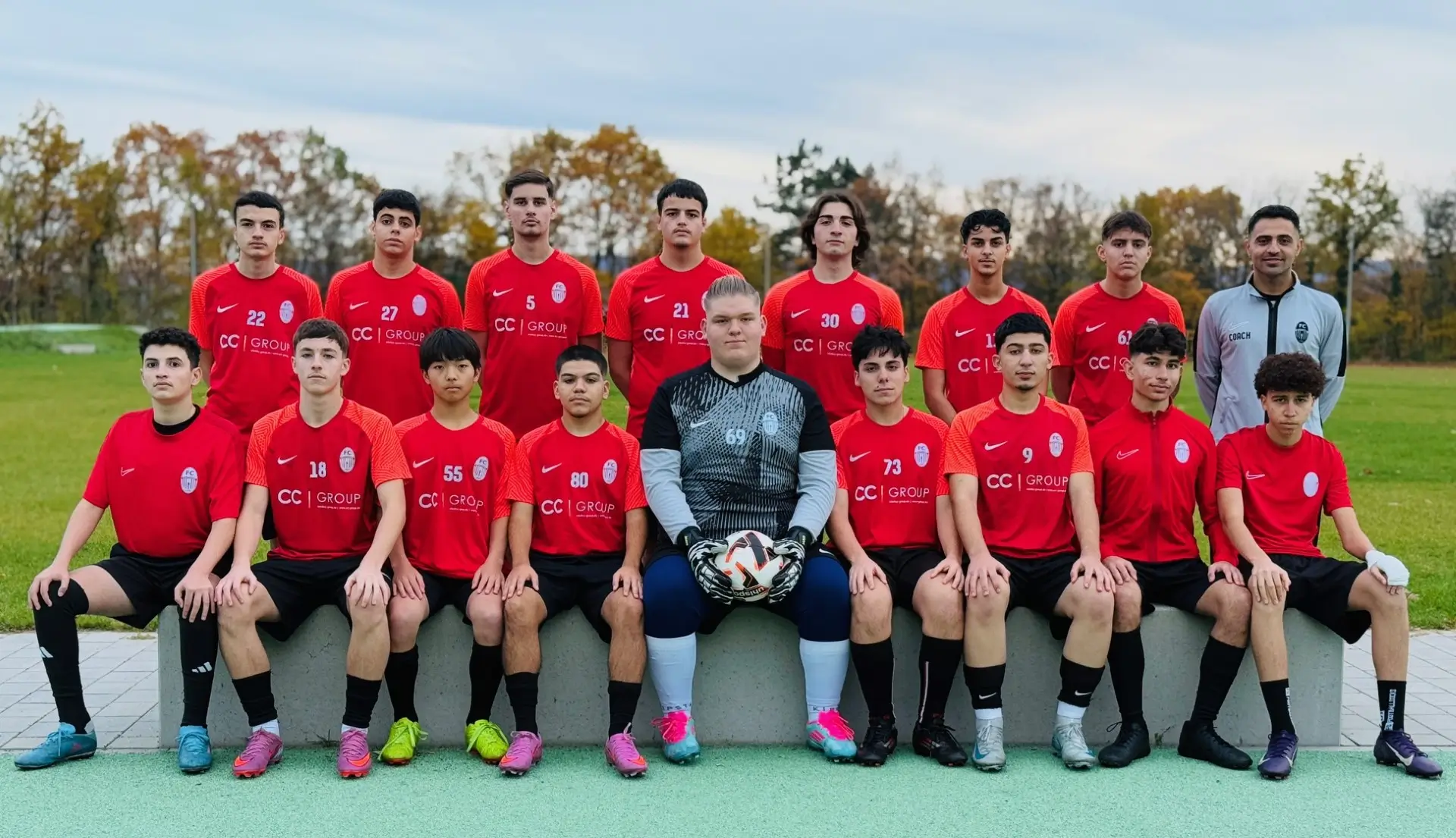 Gruppenbild einer Jugendfußballmannschaft in roten Trikots mit Torwart und Trainer auf einem Sportplatz.