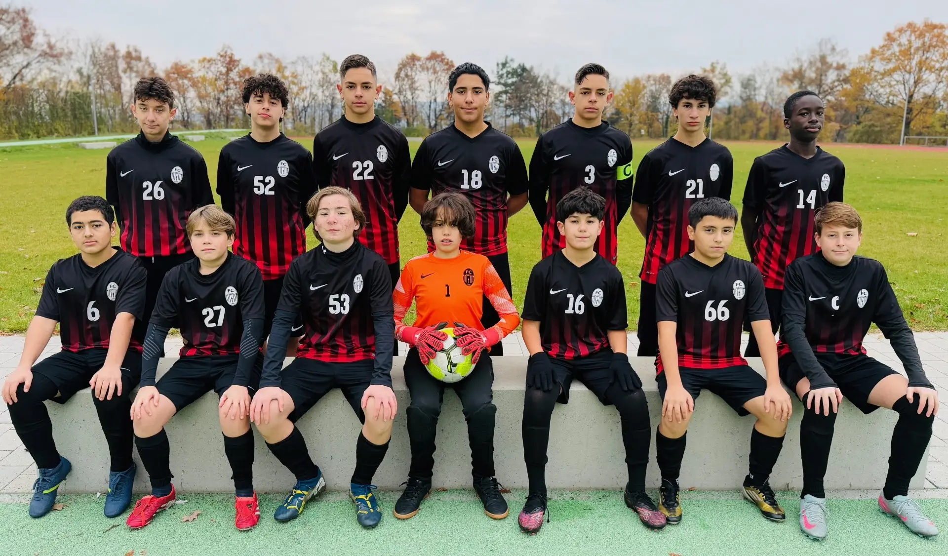 Gruppenbild einer Jugendfußballmannschaft in schwarz-roten Trikots mit Torwart auf einem Sportplatz.